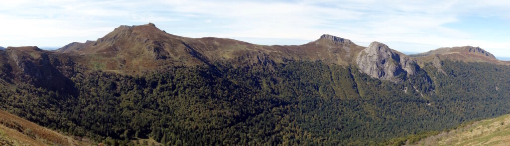 Un paysage montagneux, avec des crêtes rocheuses grises, des prairies roussies et un étage de forêts vertes en-dessous.